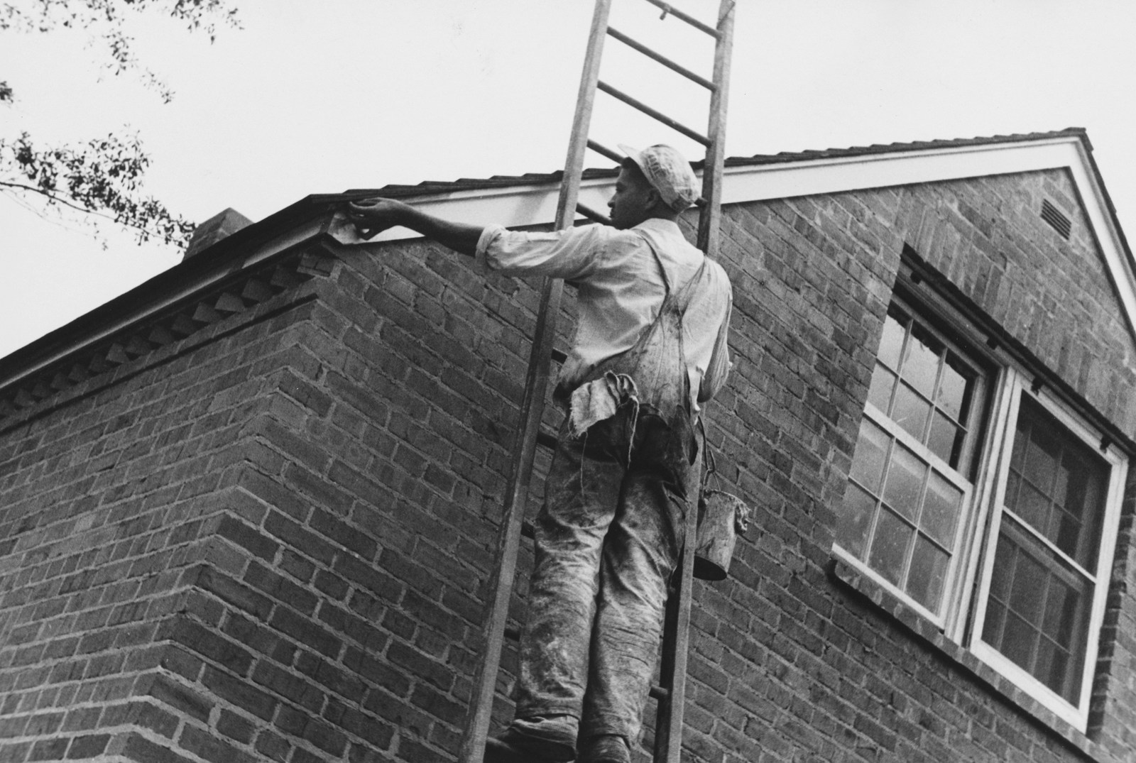 Man on ladder painting the side of a brick house.